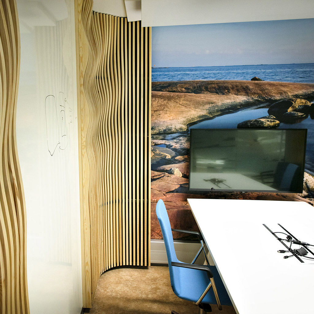 Neuvotteluhuoneen pöytä ja näyttö, aaltoilevaa puuritilää seinällä, maisemakuva toisella seinällä.