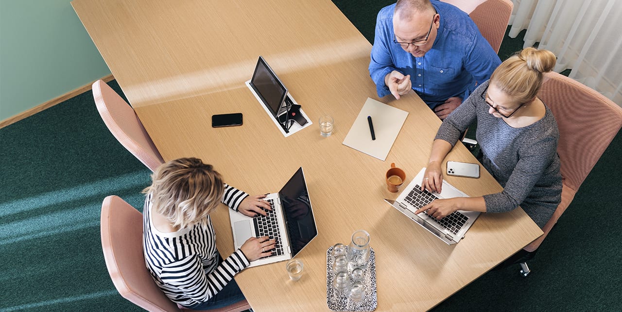 Kolme henkilöä työskentelee pöydän ääressä kannettavien tietokoneiden kanssa.
